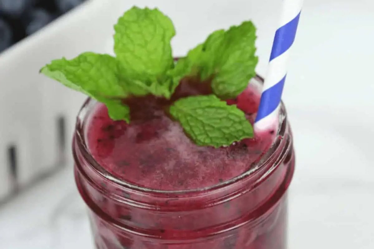 A purple blueberry moonshine in a glass jar topped with fresh mint leaves and a blue-and-white striped straw.