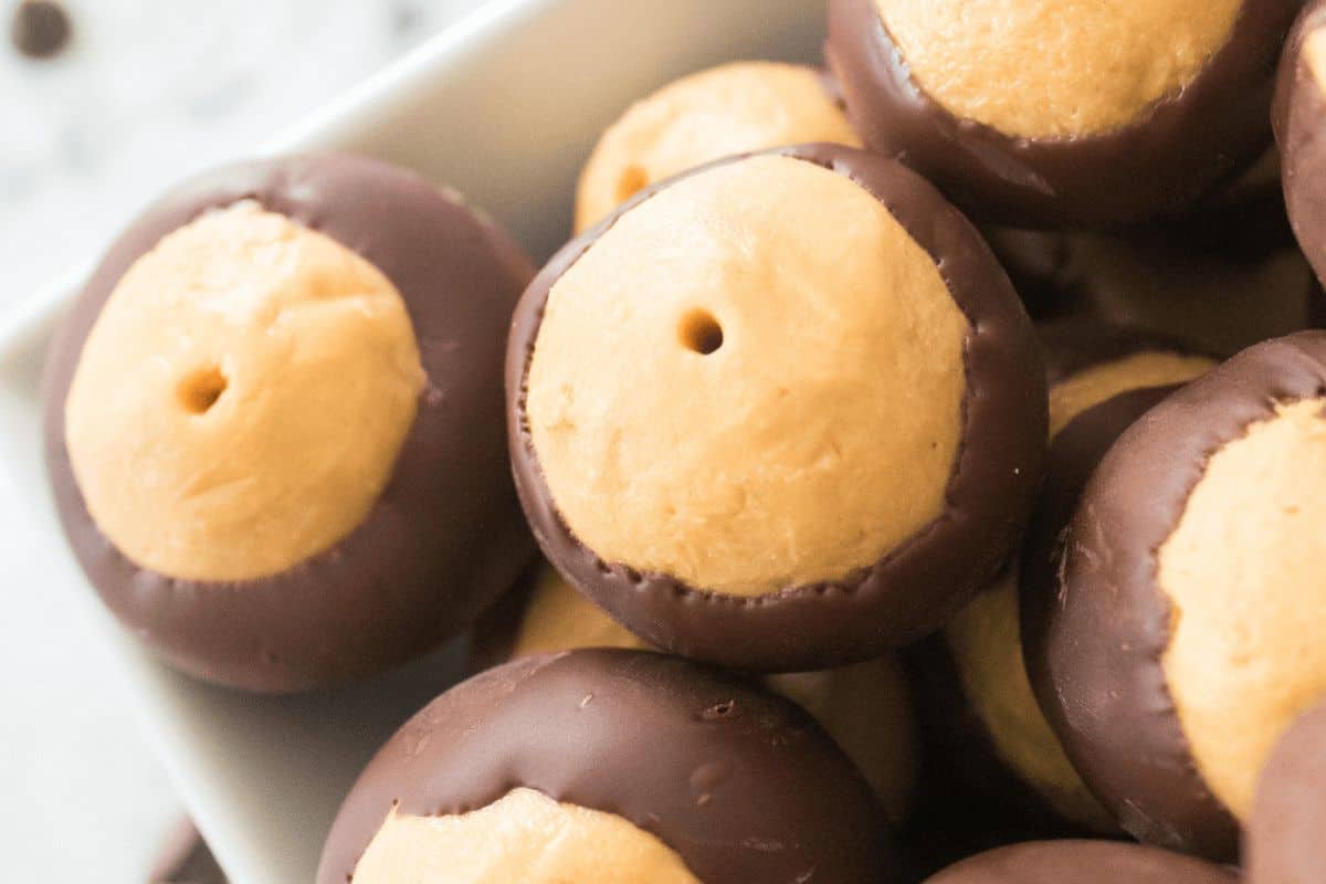 Close-up of several must-try buckeye candies. The round confections feature a smooth peanut butter center partially dipped in dark chocolate, arranged in a white dish.