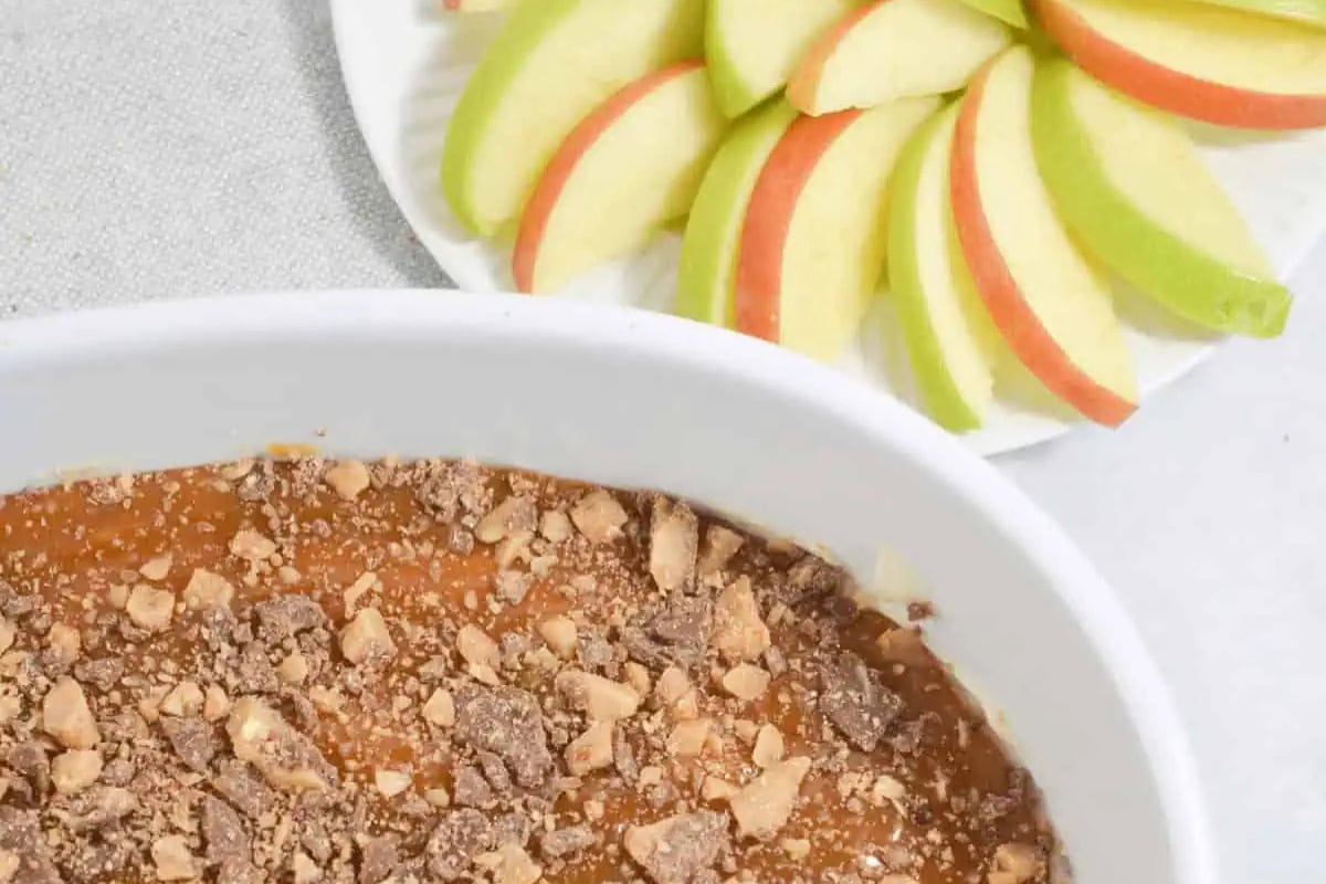 A white dish filled with caramel dip topped with crushed toffee sits next to a plate of sliced red and green apples.