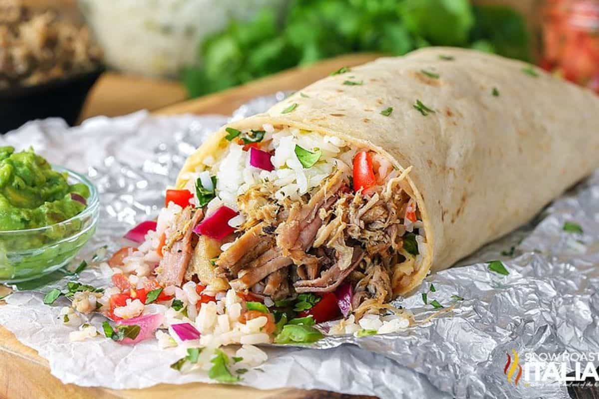 A burrito with shredded meat, rice, and vegetables on foil, perfectly paired with a small bowl of guacamole beside it. This creation tastes just like the real thing from your favorite spot.