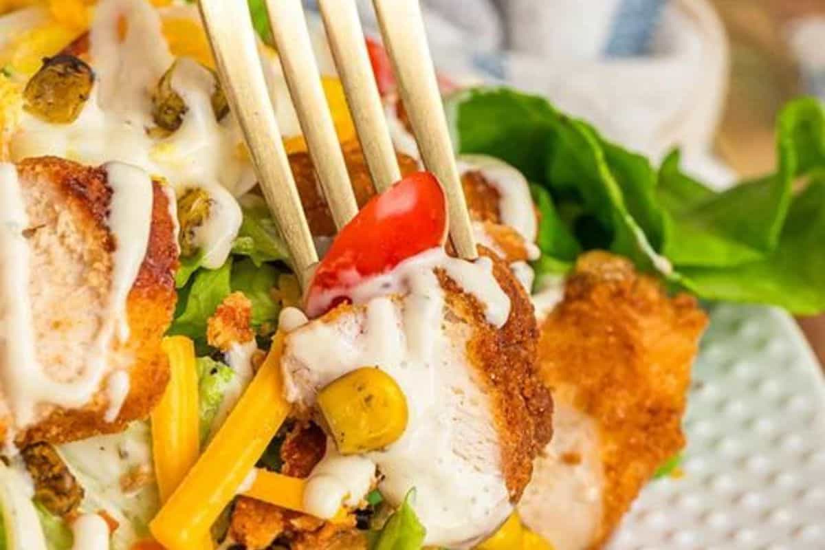 A fork lifts a piece of crispy chicken from a salad with cheese, corn, and cherry tomatoes, all topped with dressing—a dish reminiscent of Chick-fil-A recipes you can make at home.