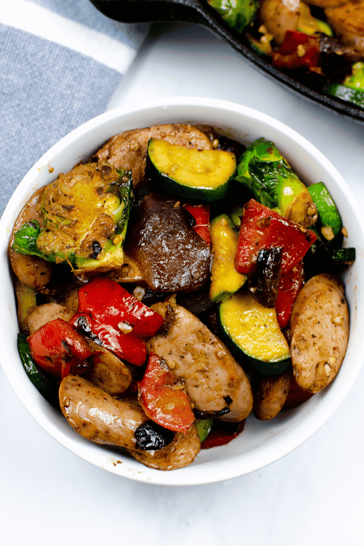 A white bowl filled with cooked apple sausage chicken slices, zucchini, red bell peppers, mushrooms, and Brussels sprouts.