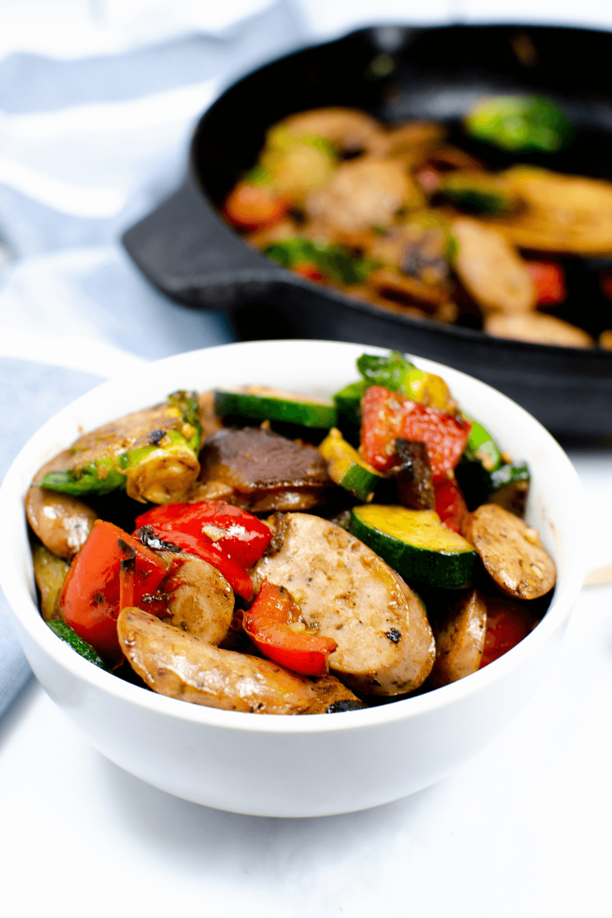 A white bowl filled with sliced apple sausage, zucchini, bell peppers, and mushrooms, with a cast iron skillet of this delicious Apple Sausage Chicken skillet dish in the background.