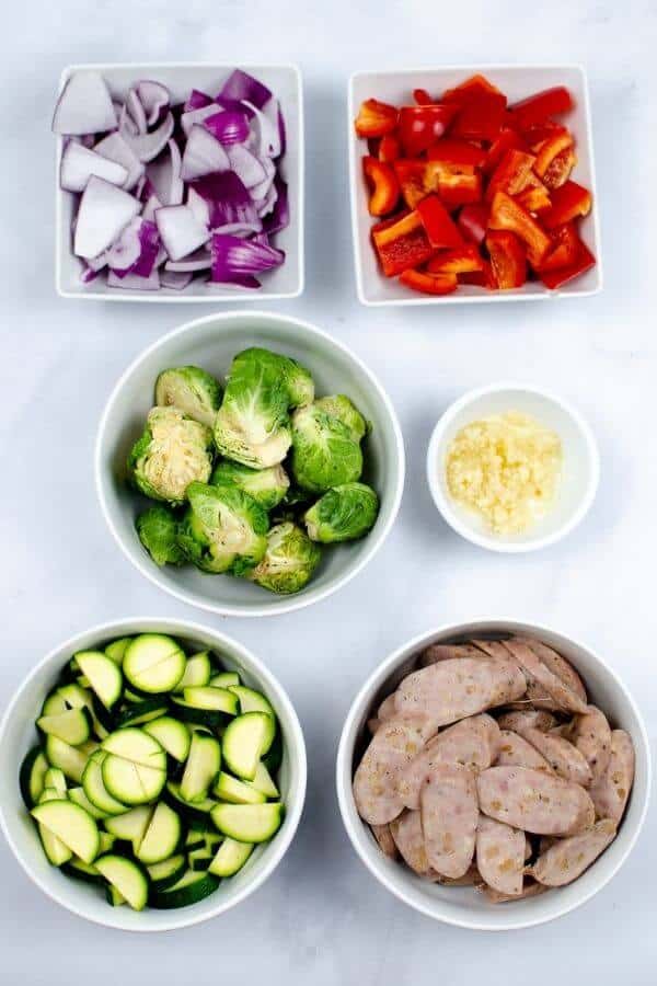 Five bowls containing chopped red onion, red bell pepper, brussels sprouts, zucchini, minced garlic, and sliced sausage—perfectly prepped for creating a vibrant Apple Sausage Chicken Skillet—are arranged on a white surface.