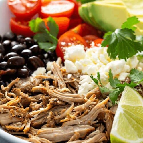A bowl inspired by Chipotle recipes, featuring shredded meat, black beans, cherry tomatoes, avocado slices, rice, queso fresco, cilantro, and lime wedges for that real thing taste.