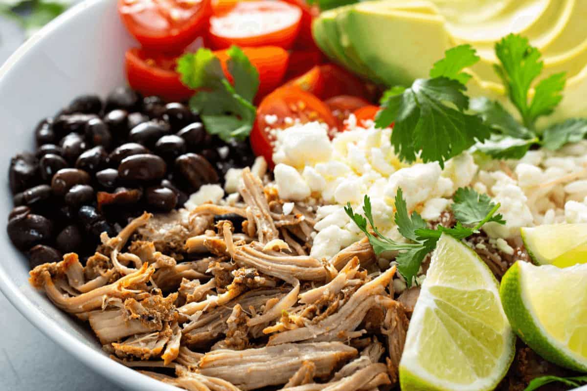 A bowl inspired by Chipotle recipes, featuring shredded meat, black beans, cherry tomatoes, avocado slices, rice, queso fresco, cilantro, and lime wedges for that real thing taste.
