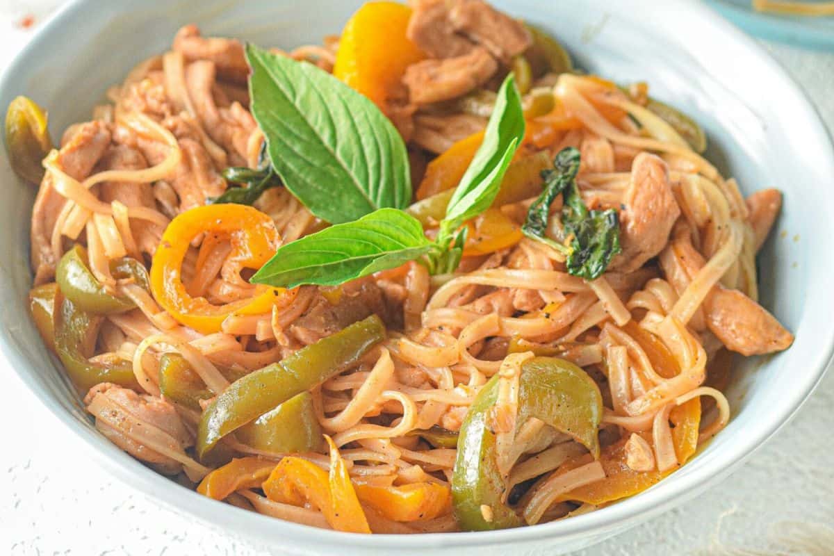 A bowl of stir-fried noodles with sliced bell peppers, pieces of meat, and fresh basil leaves as garnish—this must-try dish brings the bold flavors of Asian dishes right to your table, better than takeout.