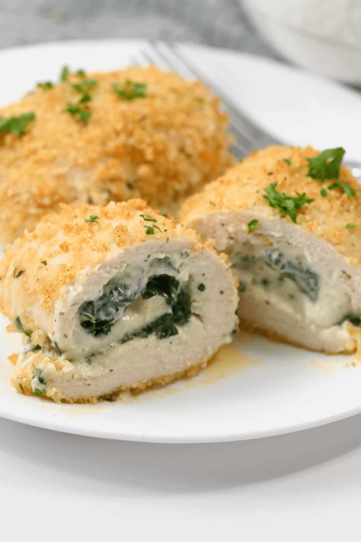 A plate with sliced Chicken Rollatini, featuring creamy spinach filling and a crispy breadcrumb coating, garnished with chopped herbs.
