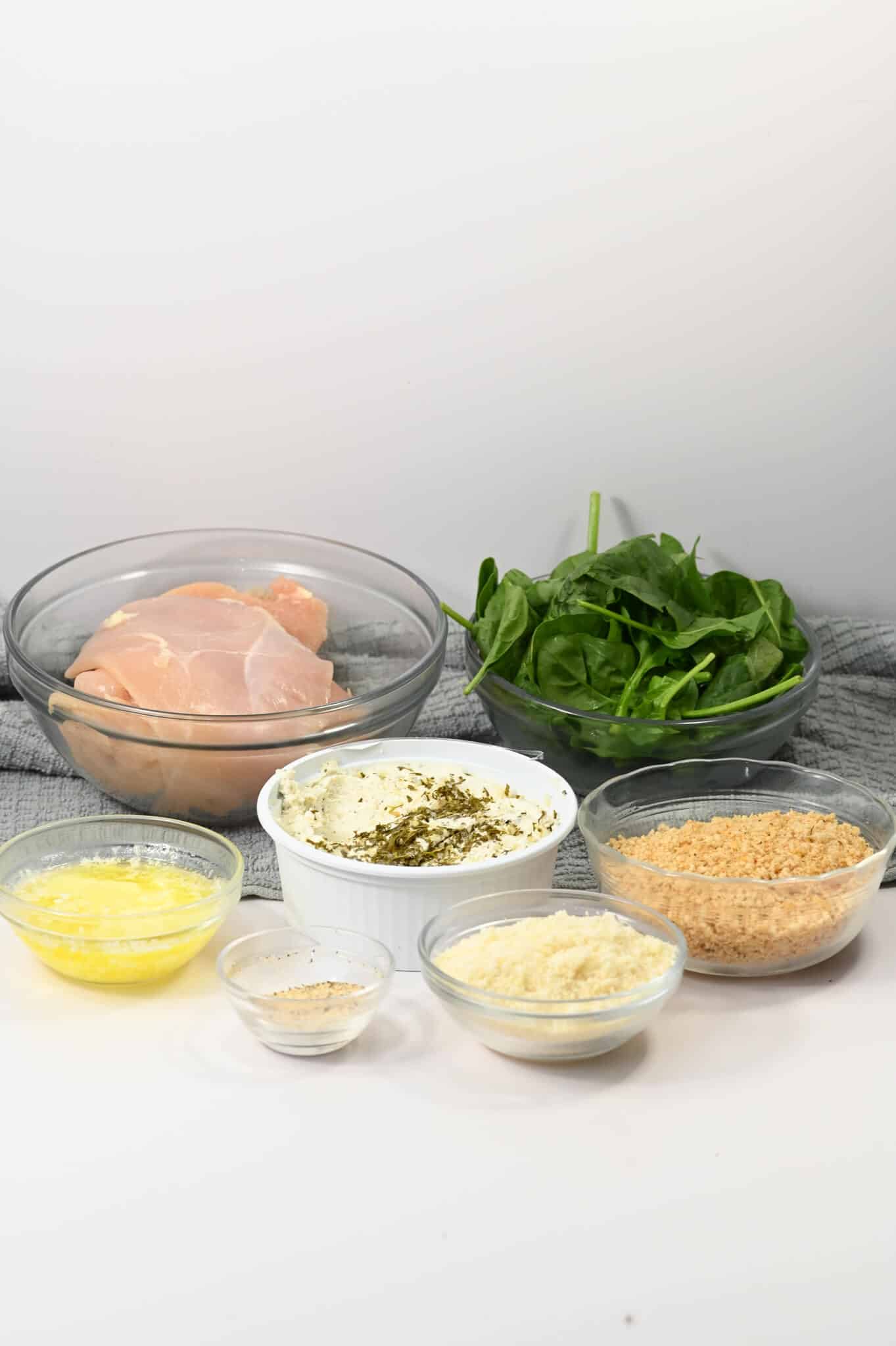 Displayed on a gray towel against a white background are various ingredients raw chicken, spinach, cream cheese with herbs, breadcrumbs, shredded cheese, melted butter, and seasonings. Each component is artfully arranged in bowls to capture the essence of this delicious dish.