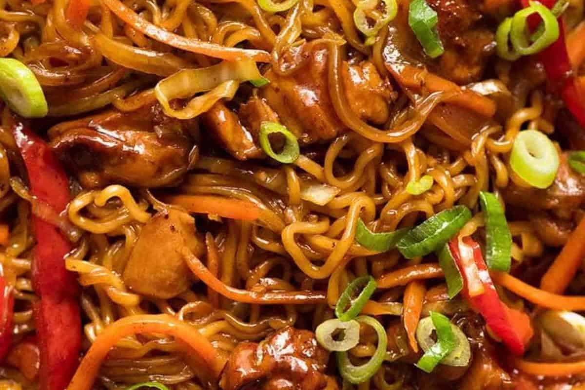 Close-up of stir-fried noodles with sliced carrots, red bell peppers, green onions, and pieces of chicken in a savory sauce—a must try for fans of Asian dishes.