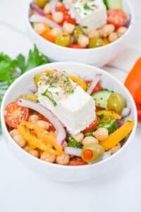 A bowl of Greek Chickpea Salad with cherry tomatoes, olives, bell peppers, red onion, cucumber, parsley, and a slice of feta cheese on top.