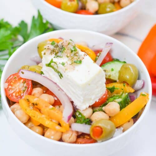 A bowl of Greek Chickpea Salad with cherry tomatoes, olives, bell peppers, red onion, cucumber, parsley, and a slice of feta cheese on top.