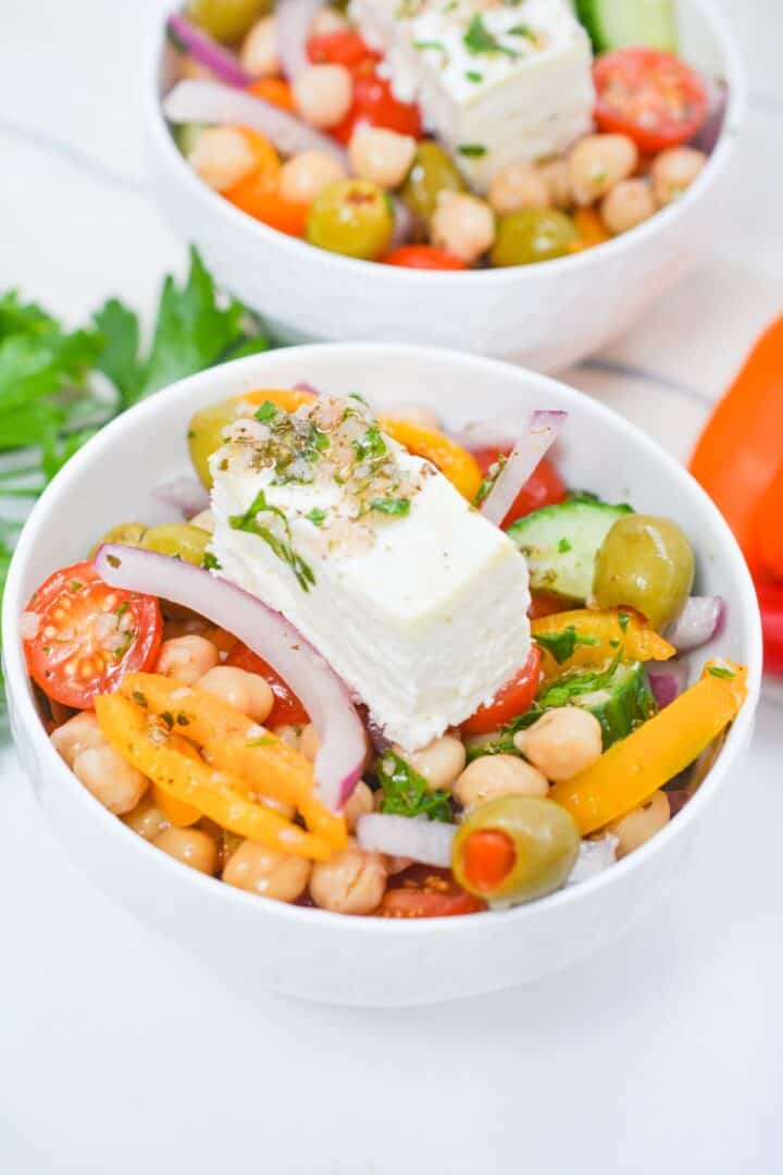 A bowl of Greek Chickpea Salad with cherry tomatoes, olives, bell peppers, red onion, cucumber, parsley, and a slice of feta cheese on top.