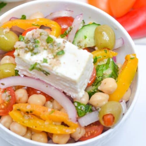 A vibrant Greek Chickpea Salad is served in a bowl, featuring chickpeas, cherry tomatoes, olives, cucumber, yellow bell pepper, red onion, fresh herbs, and a block of feta cheese. Parsley and tomatoes are arranged on the side for an extra burst of flavor and color.