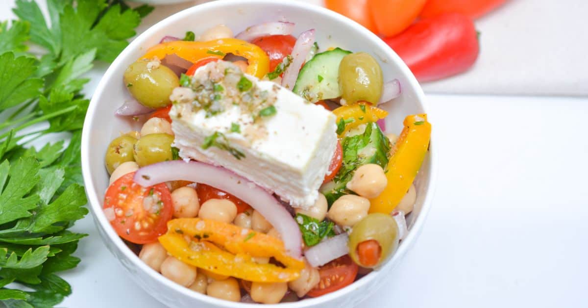 A vibrant Greek Chickpea Salad is served in a bowl, featuring chickpeas, cherry tomatoes, olives, cucumber, yellow bell pepper, red onion, fresh herbs, and a block of feta cheese. Parsley and tomatoes are arranged on the side for an extra burst of flavor and color.