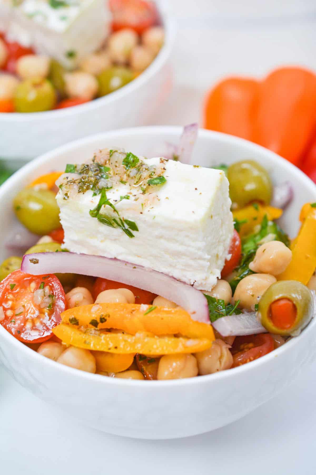 A bowl of Greek Chickpea Salad with cherry tomatoes, yellow peppers, red onion, green olives, and a slice of feta cheese topped with herbs.