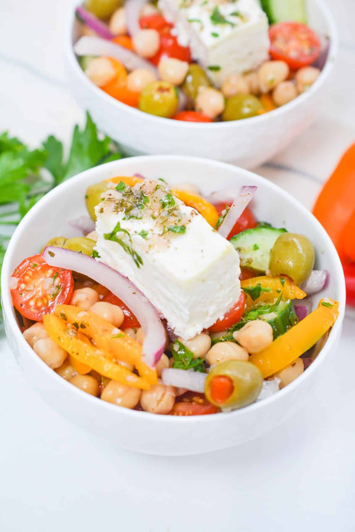 A bowl of Greek Chickpea Salad featuring sliced peppers, cherry tomatoes, red onion, green olives, and a block of feta cheese on top.