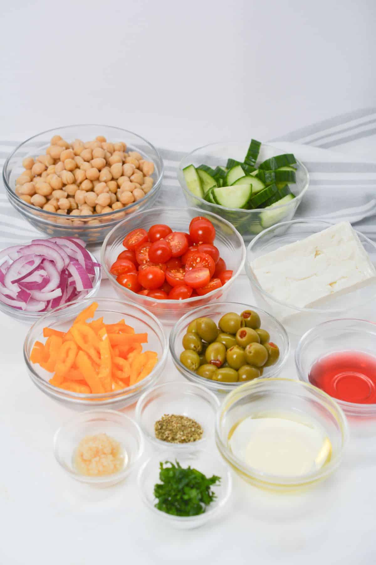 Assorted Greek Chickpea Salad ingredients in bowls, including chickpeas, cucumber, cherry tomatoes, red onion, bell pepper, feta cheese, green olives, olive oil, red wine vinegar, herbs, and garlic.