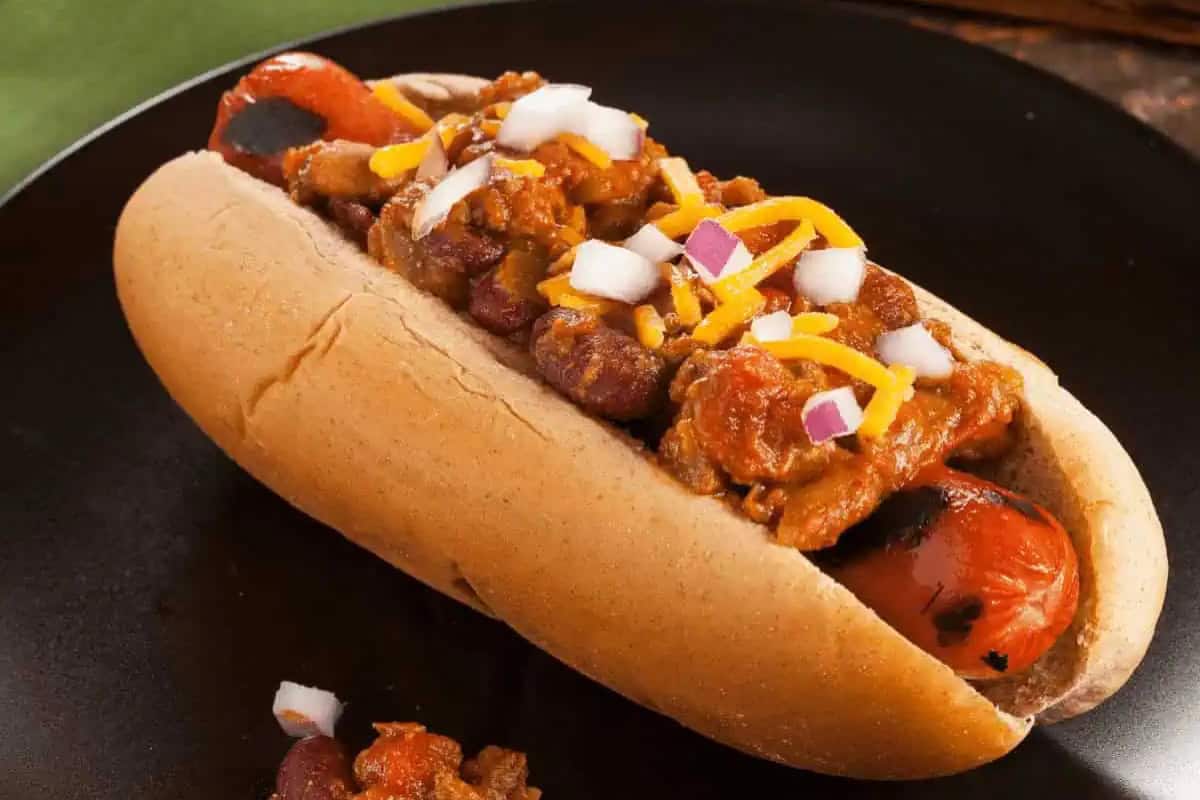 A grilled hot dog in a bun topped with chili, kidney beans, shredded cheddar cheese, and chopped onions, served on a dark plate.