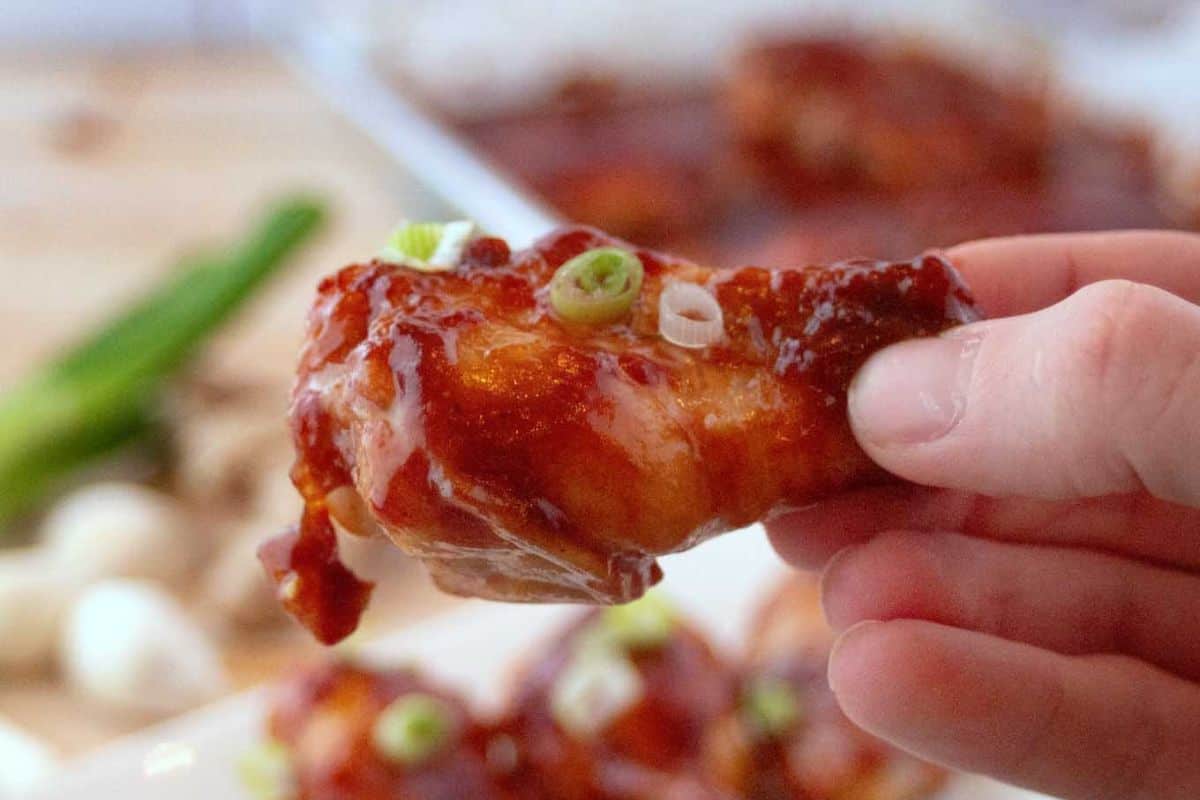 A hand holds a glazed chicken wing garnished with sliced green onions, with more wings and green onions in the background—this must-try dish is perfect for fans of Asian dishes.