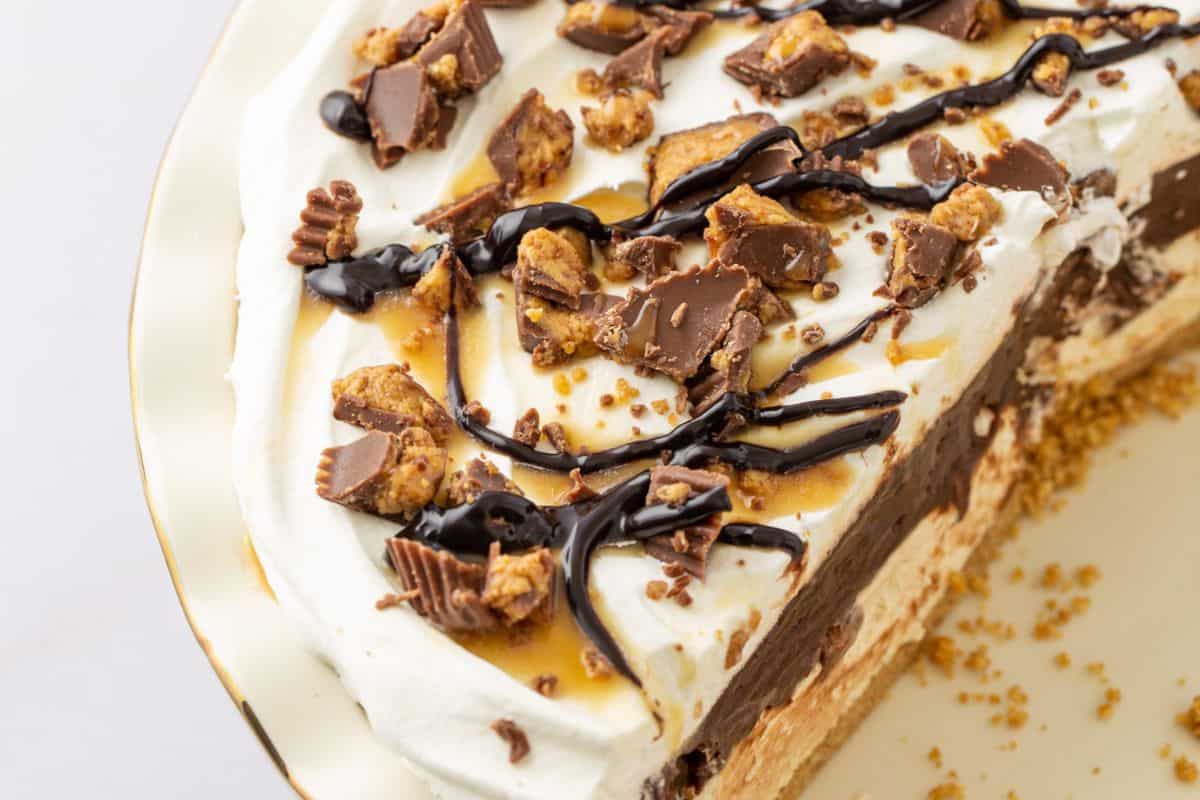 A close-up of a slice of pie with a graham cracker crust, layers of chocolate and peanut butter fillings, topped with whipped cream, chocolate syrup, caramel drizzle, and chopped peanut butter cups—this is one of those must-try peanut butter recipes that's simply irresistible.