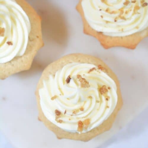 Two cinnamon cupcakes with white frosting and sprinkled nuts on top sit on a light surface, with part of a third cupcake visible in the background.