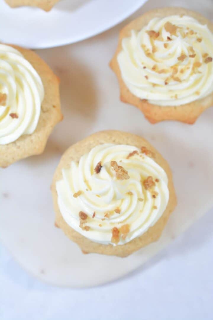 Two cinnamon cupcakes with white frosting and sprinkled nuts on top sit on a light surface, with part of a third cupcake visible in the background.