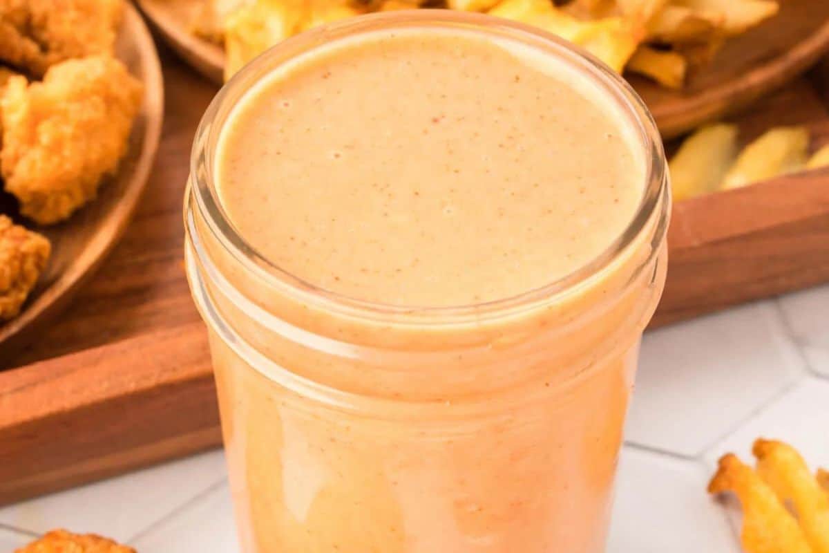 A jar filled with creamy dipping sauce is placed on a table, reminiscent of Chick-fil-A recipes. In the background, there are fries and breaded snacks on a wooden tray. Perfect for those looking to make at home delights that capture the essence of secret recipes.