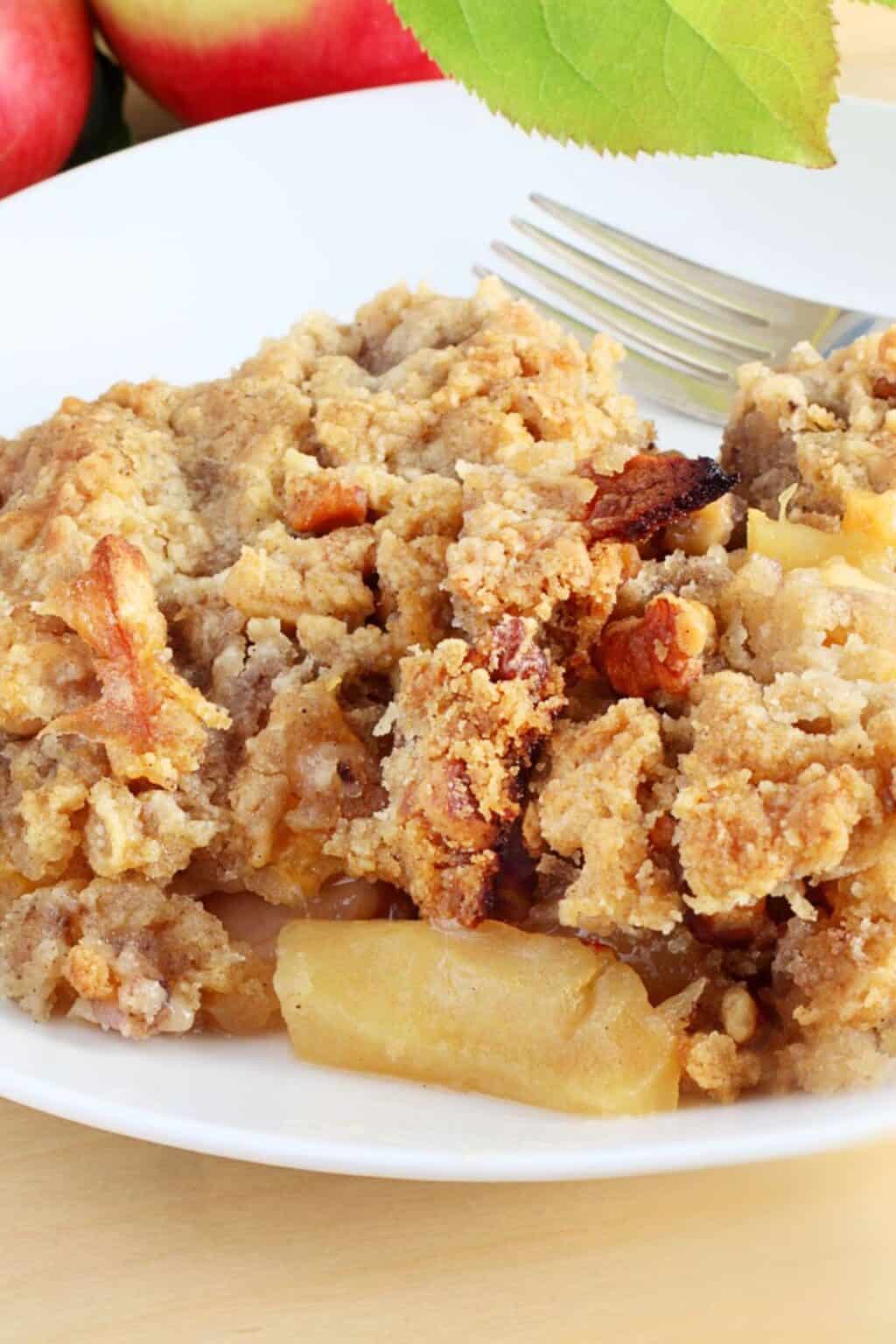 A serving of crock pot apple crisp with a crumbly topping sits on a white plate, accompanied by a fork. Some red apples and a green leaf are in the background.
