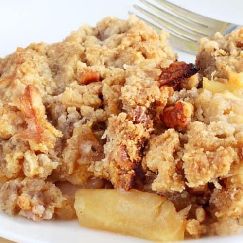 A serving of crock pot apple crisp with a crumbly topping sits on a white plate, accompanied by a fork. Some red apples and a green leaf are in the background.