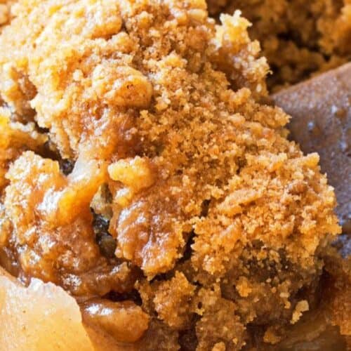 Close-up of a baked Apple Crisp dessert with a golden-brown, crumbly topping and visible apple slices, next to a wooden spoon. Perfect for making in your Crock Pot for an easy and delicious treat.