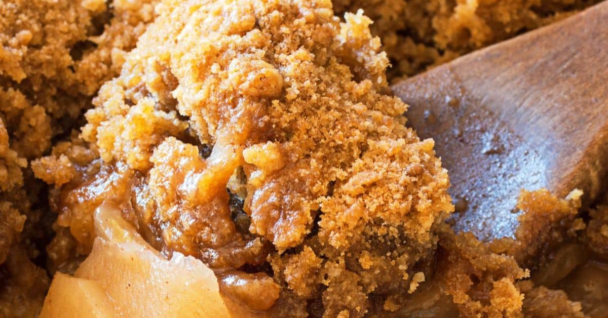 Close-up of a baked Apple Crisp dessert with a golden-brown, crumbly topping and visible apple slices, next to a wooden spoon. Perfect for making in your Crock Pot for an easy and delicious treat.