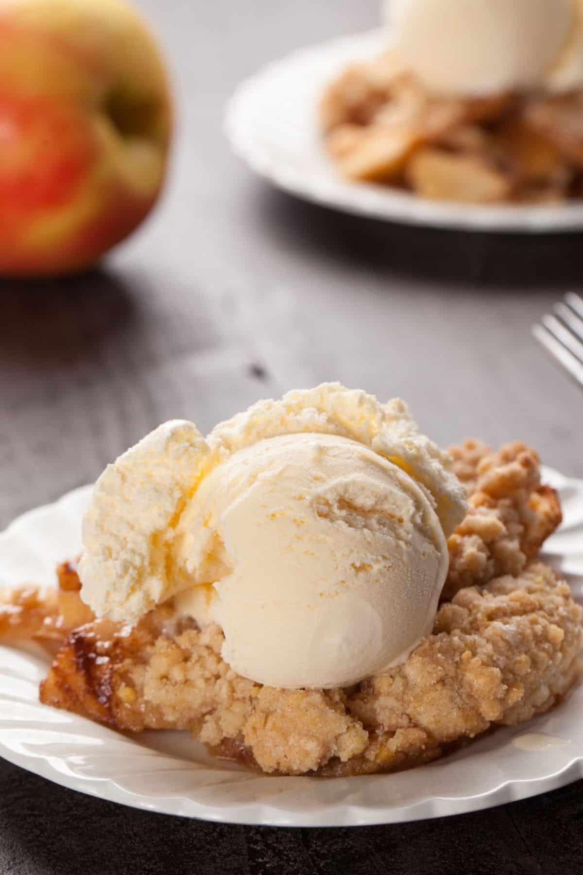A slice of apple crumble pie topped with a scoop of vanilla ice cream on a white plate, reminiscent of the comforting flavors found in Crock Pot Apple Crisp, with an apple and another dessert plate in the background.