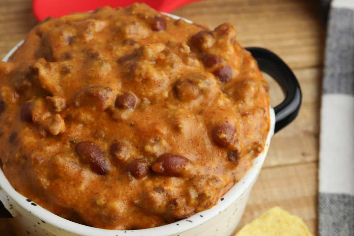 A close-up of a bowl filled with thick, cheesy chili dip containing ground meat and beans, set on a wooden surface.