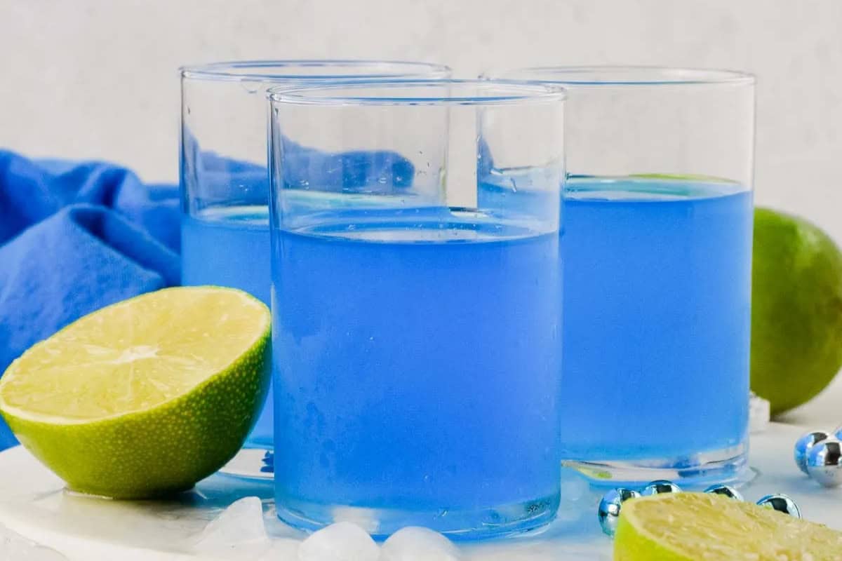 Three clear glasses filled with blue Kool-Aid cocktails sit on a white surface, surrounded by lime halves, ice cubes, and silver decorative beads.
