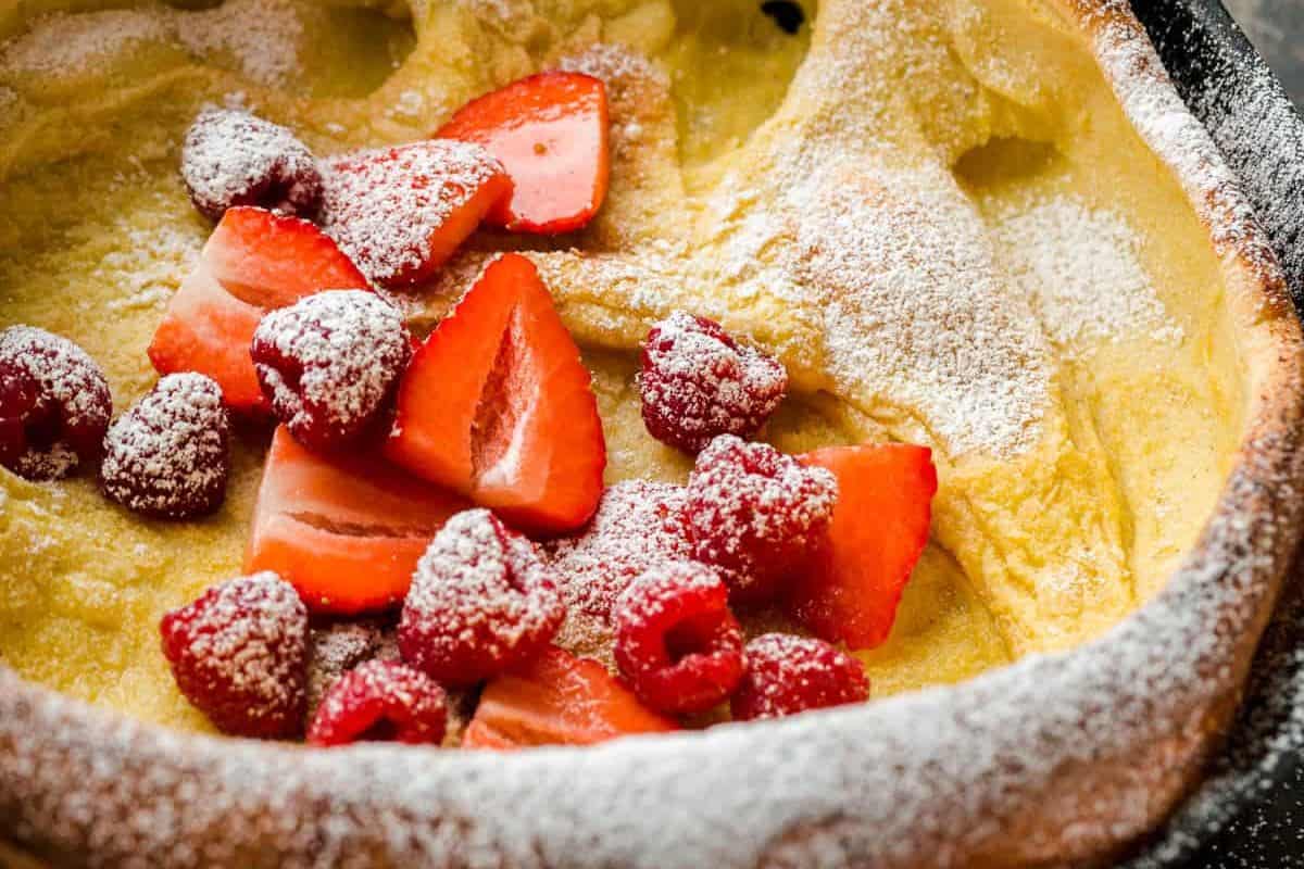 A close-up of a Dutch baby pancake topped with sliced strawberries, raspberries, and a dusting of powdered sugar, perfect for those who make every weekend special with delicious pancake recipes.