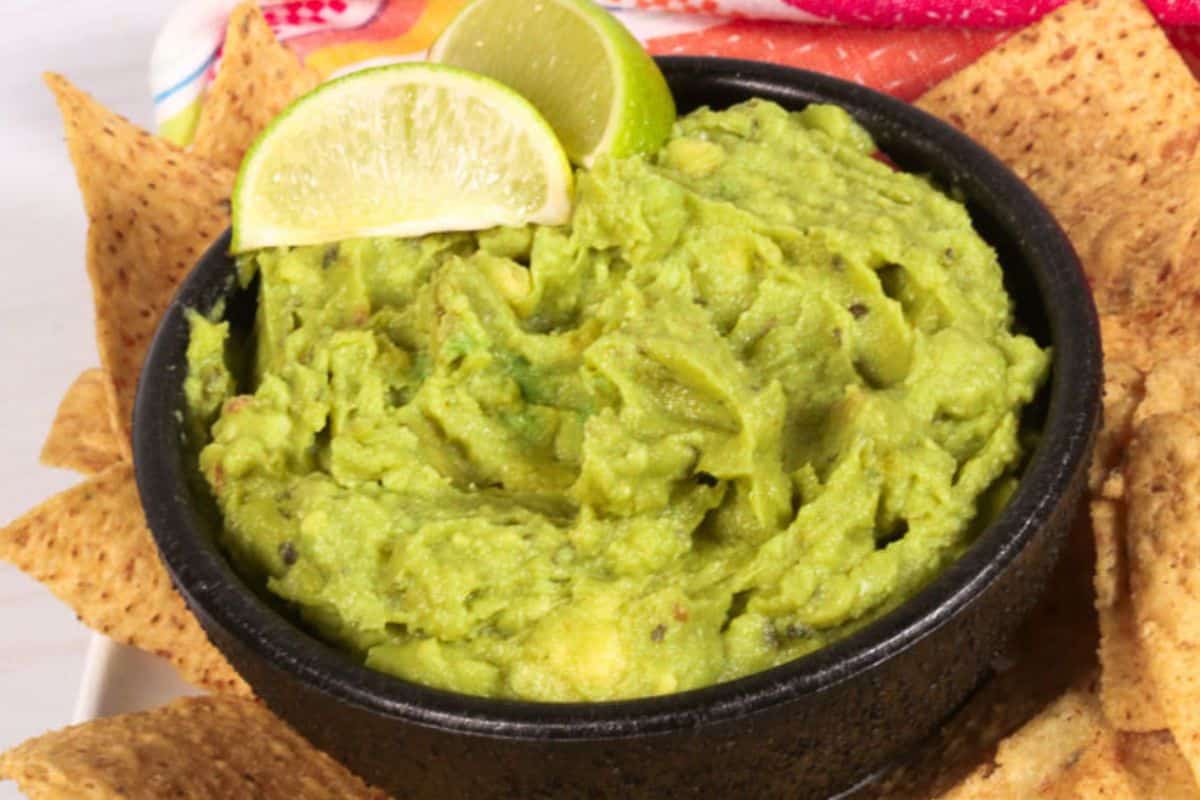 A black bowl of guacamole, crafted as a copycat Chipotle recipe, is topped with lime slices and surrounded by crunchy tortilla chips.
