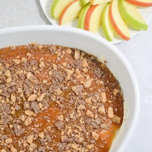 A white dish with rich caramel dip, toffee-topped for extra sweetness, sits next to a plate of sliced red and green apples arranged in a fan shape.