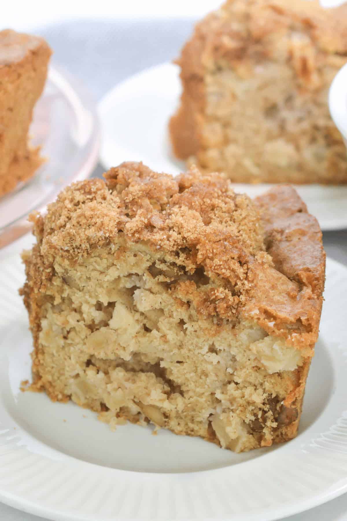 A slice of crumb-topped Fresh Apple Cake, with visible apple chunks, sits on a white plate, with more cake in the background.