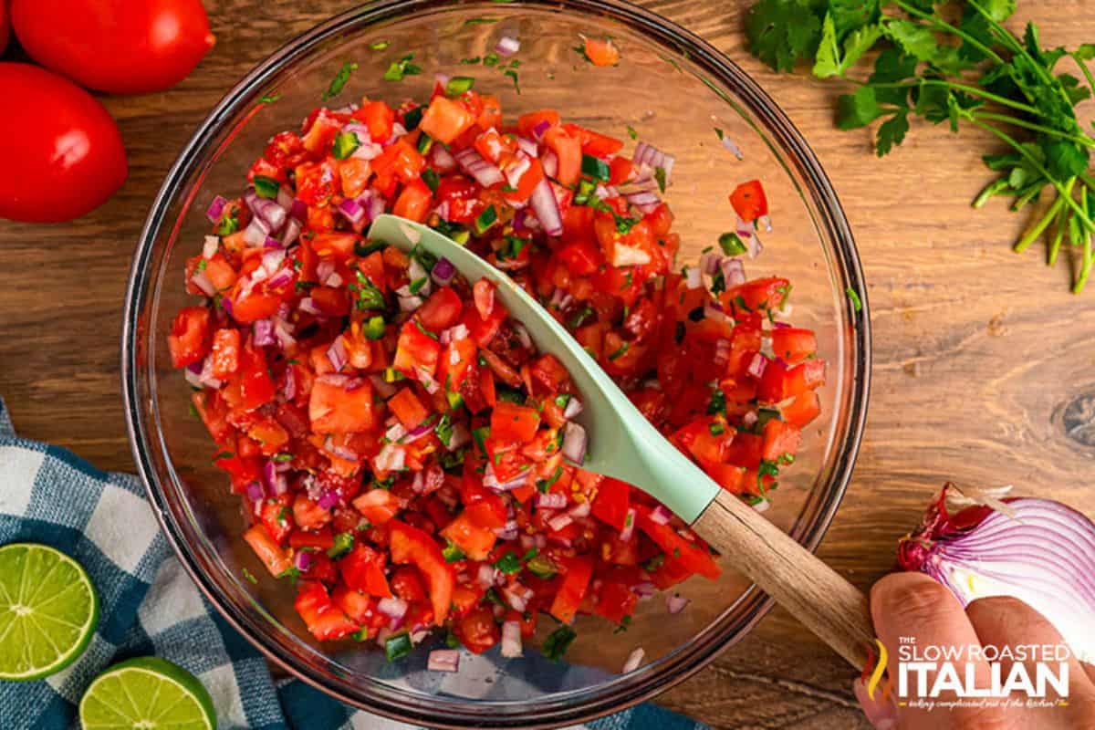 A bowl of fresh salsa with diced tomatoes, onions, and herbs, inspired by Real Thing Recipes, is accompanied by a spatula. Fresh tomatoes, a red onion, lime halves, and cilantro are placed nearby on a wooden surface.