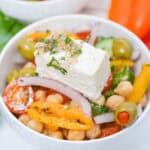 A bowl of Greek Chickpea Salad with cherry tomatoes, sliced bell peppers, red onion, olives, cucumber, and a block of feta cheese, garnished with herbs.