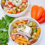 Two bowls feature cherry tomatoes, olives, bell peppers, red onion, cucumber, and feta, garnished with herbs. Fresh parsley and mini bell peppers are on the side.