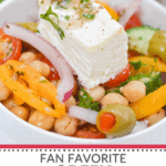 A vibrant Greek Chickpea Salad, featuring a medley of feta, peppers, onion, tomatoes, cucumber, and olives, topped with fresh herbs.