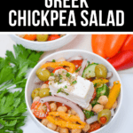 A vibrant bowl of Greek Chickpea Salad filled with olives, cherry tomatoes, cucumber, and red onion topped with a block of feta. It's elegantly garnished with parsley and accompanied by small red peppers on the side.