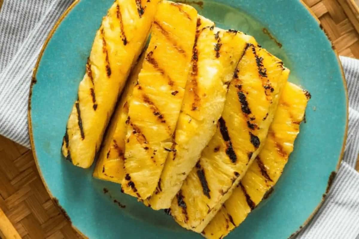 Grilled pineapple slices with char marks are arranged on a turquoise plate with a gray and white striped towel underneath. Underneath the towel is a woven wooden table.