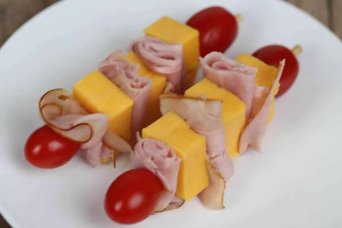 Skewers with rolled deli ham, cheddar cheese cubes, and cherry tomatoes are arranged on a white plate.
