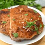 Two Honey Garlic Pork Chops, grilled to perfection and garnished with fresh herbs, lie on a white plate set on a wooden cutting board, accompanied by a bunch of leafy greens in the background.