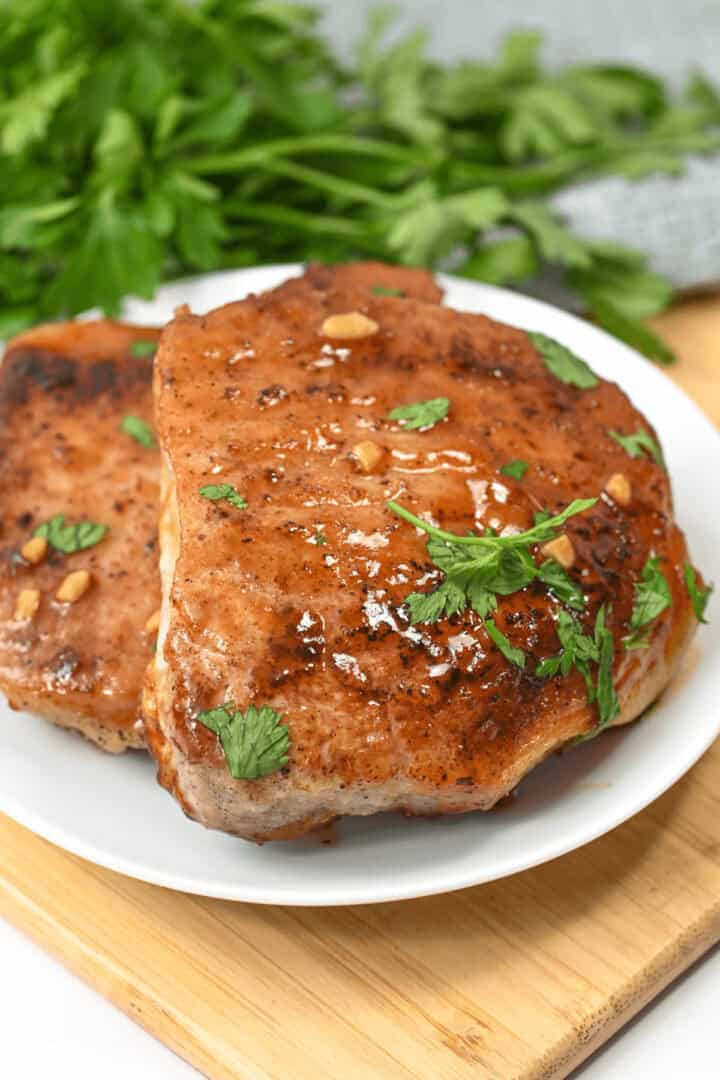 Two Honey Garlic Pork Chops, grilled to perfection and garnished with fresh herbs, lie on a white plate set on a wooden cutting board, accompanied by a bunch of leafy greens in the background.