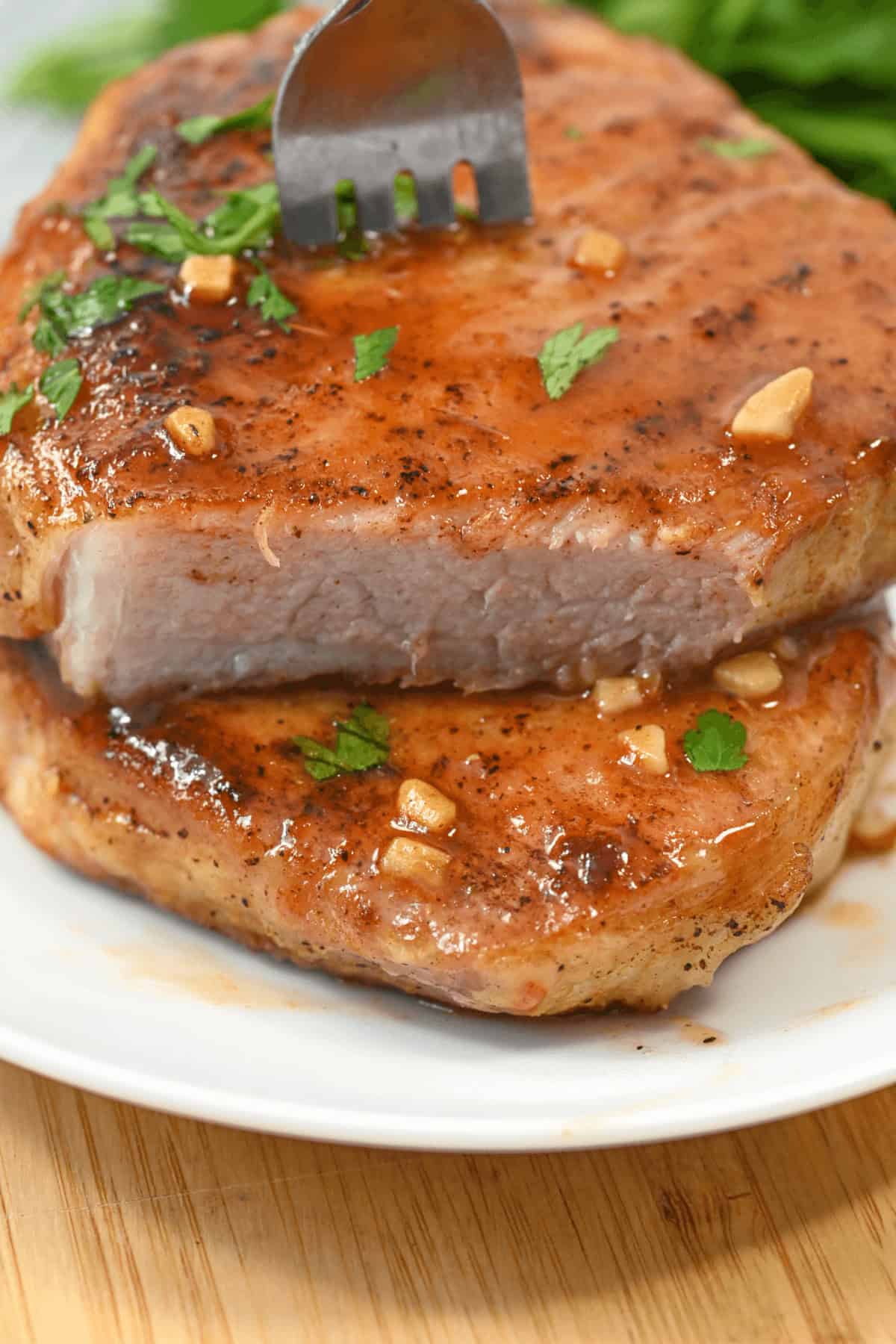 A fork pierces a juicy, cooked pork chop with garlic and herbs on a white plate, perfectly capturing the essence of delicious Honey Garlic Pork Chops.