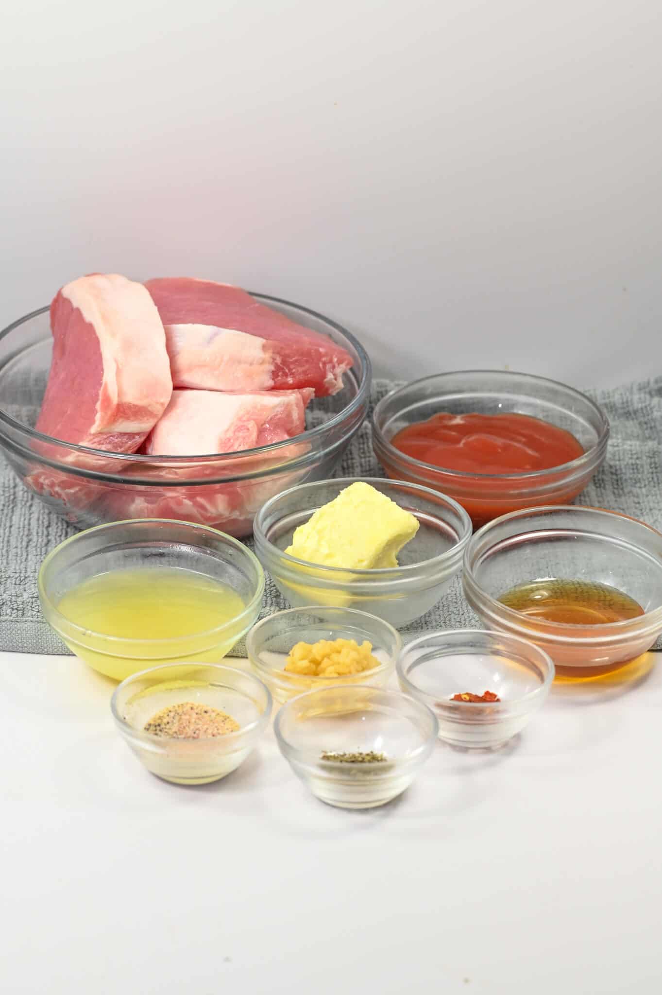 A variety of ingredients arranged in small glass bowls on a light surface, including raw pork.